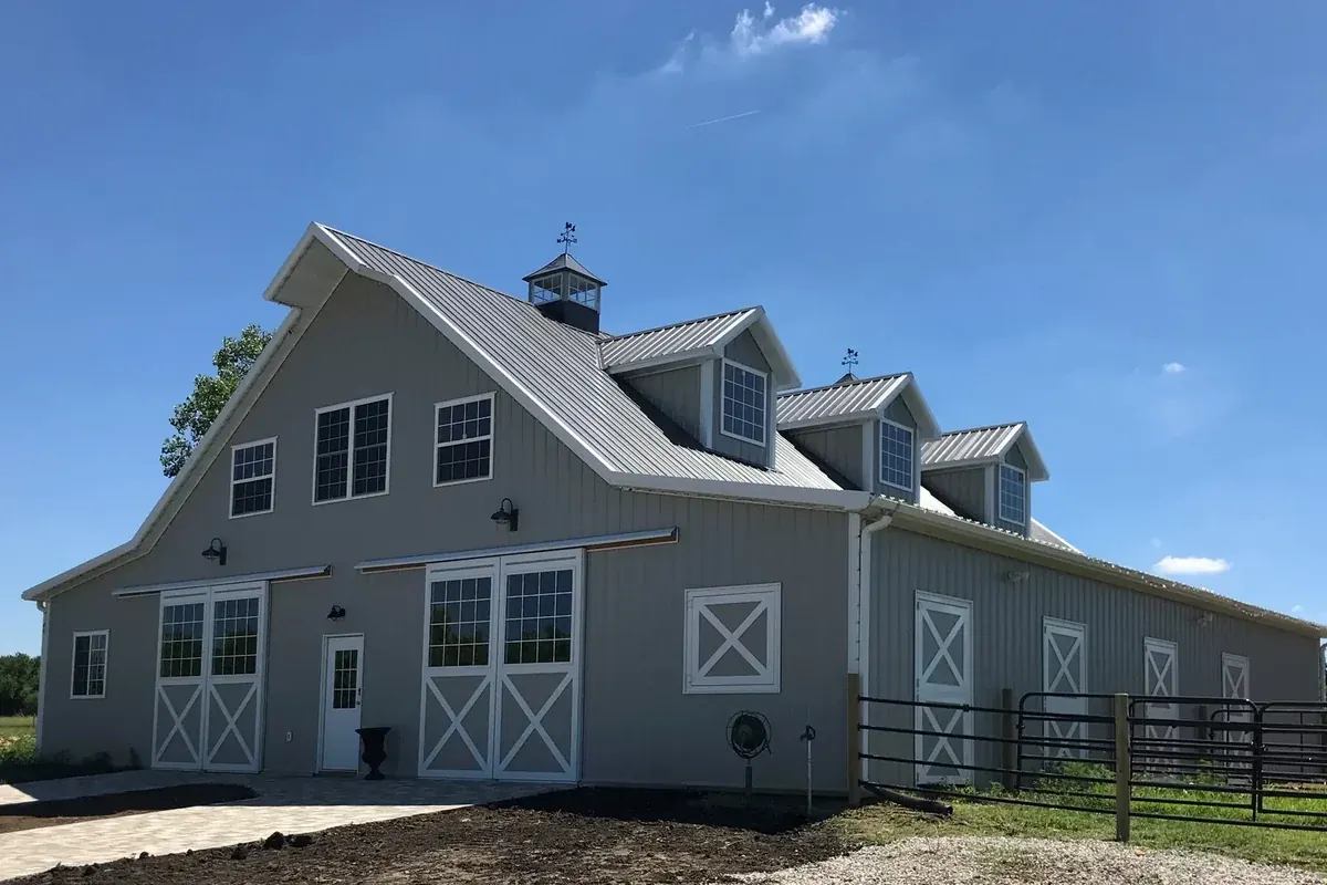 Two-story gray gambrel-roof equestrian barn with dormer windows and sliding stall doors