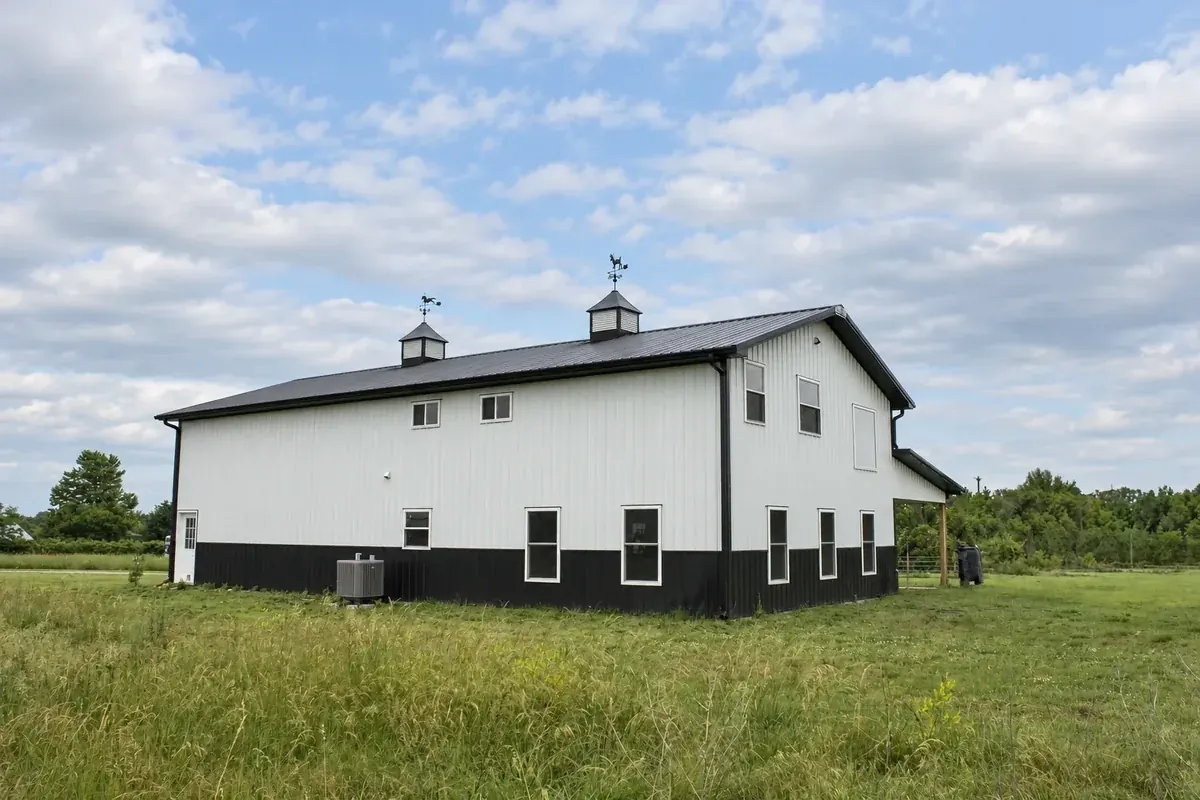 White two-story barndominium with twin roof cupolas and an attached carport