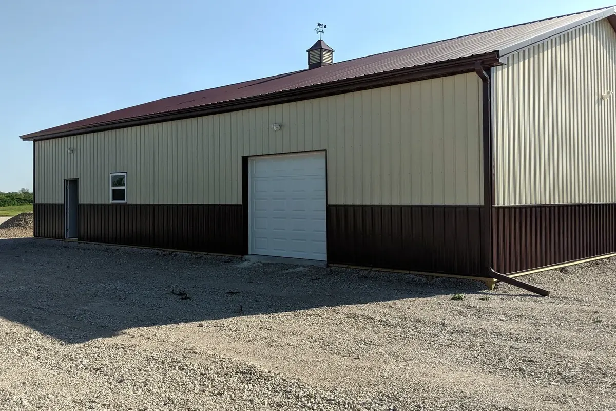 Long tan and dark maroon pole barn with a single overhead door and a roof weathervane